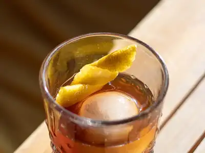 An amber stirred drink over a large round ice cube with a lemon twist