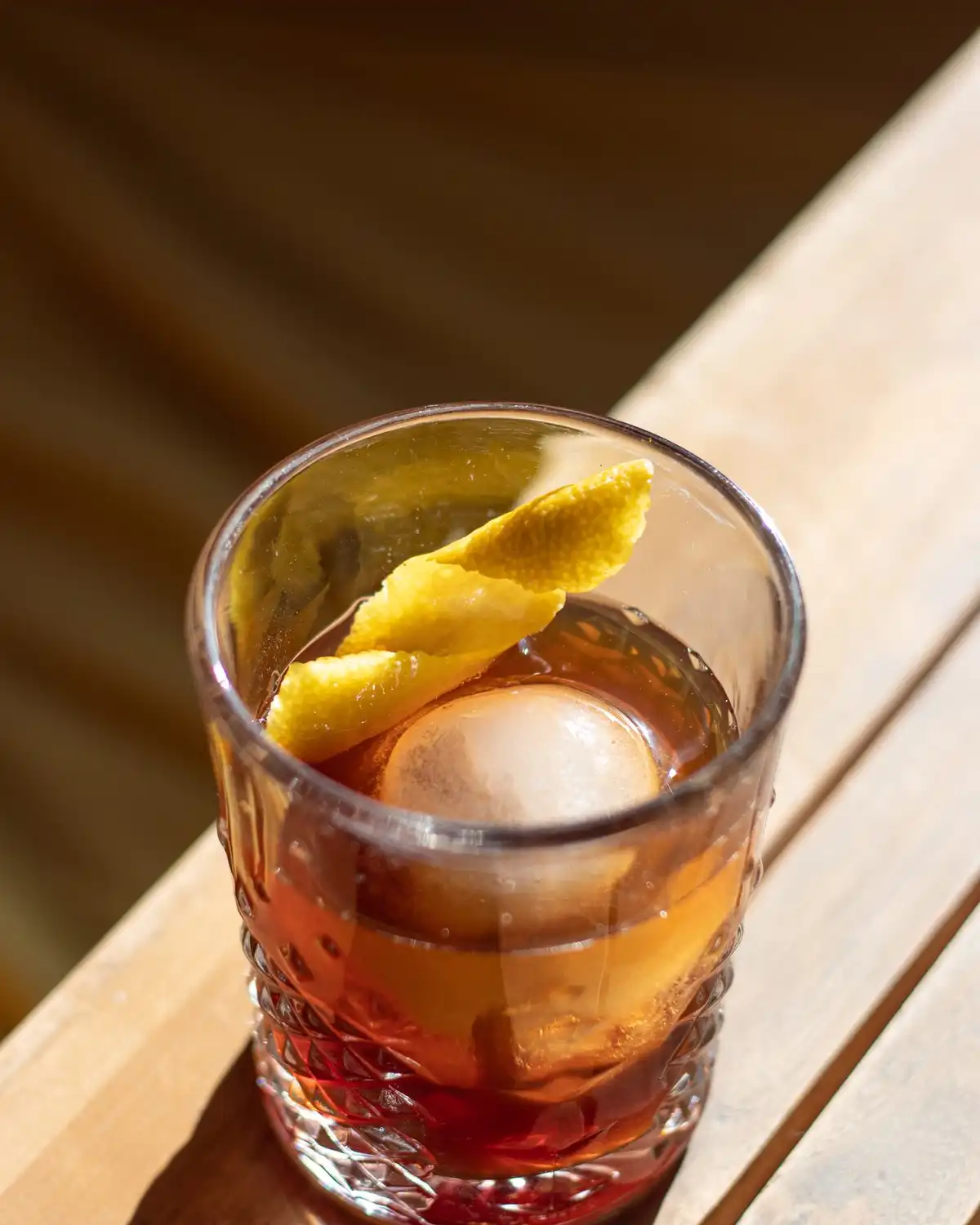 An amber stirred drink over a large round ice cube with a lemon twist