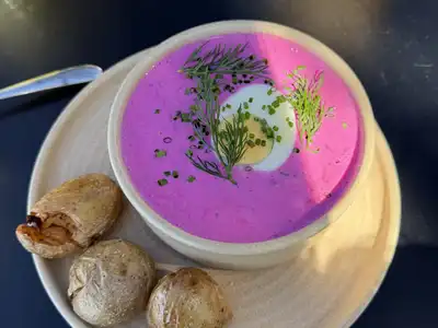 A bowl of cold pink beet soup with dill and half an egg, with small rolls on the side