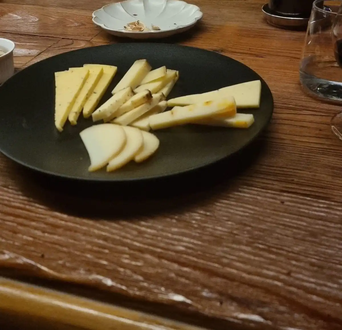 Slices of hard cheese on a black plate beside a glass of red wine on a wooden bar