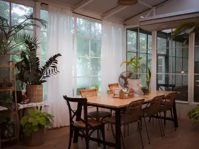 A sunny dining room with tall windows, white curtains, potted plants and a long wooden table