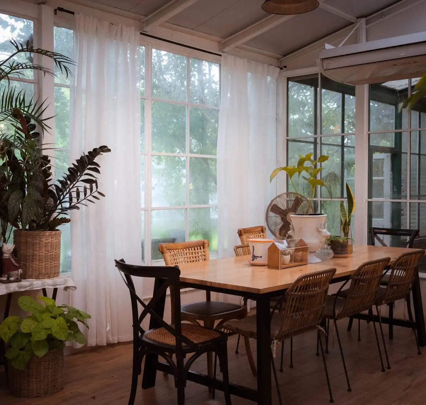 A sunny dining room with tall windows, white curtains, potted plants and a long wooden table