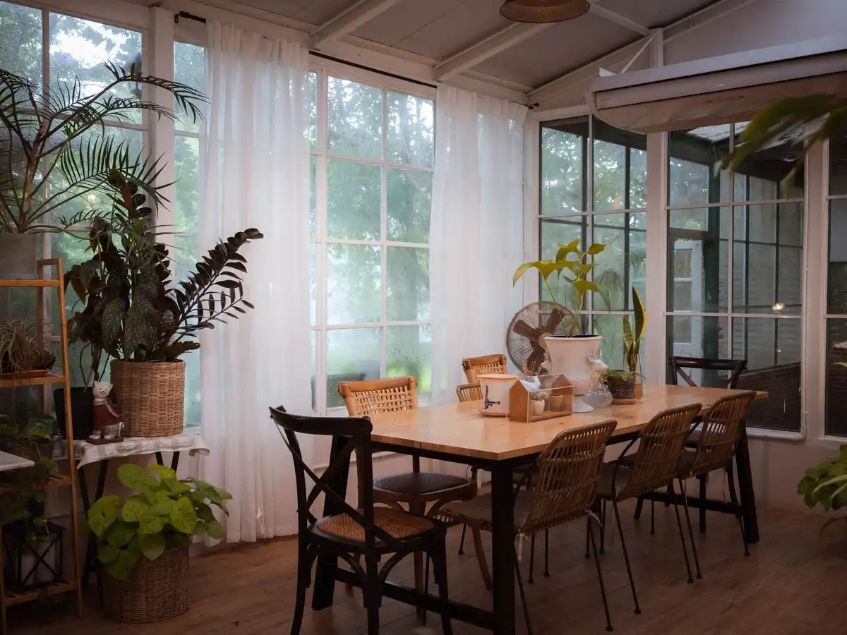 A sunny dining room with tall windows, white curtains, potted plants and a long wooden table