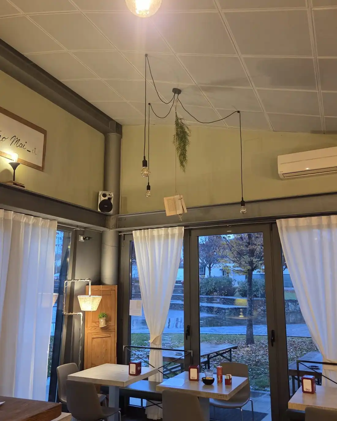 A tall dining room with a pendant light, white walls and curtained glass doors onto a park