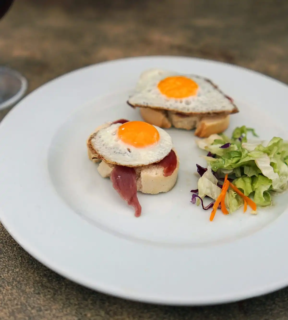 Small toasts topped with cured ham and fried quail eggs, with a side salad, on a white plate
