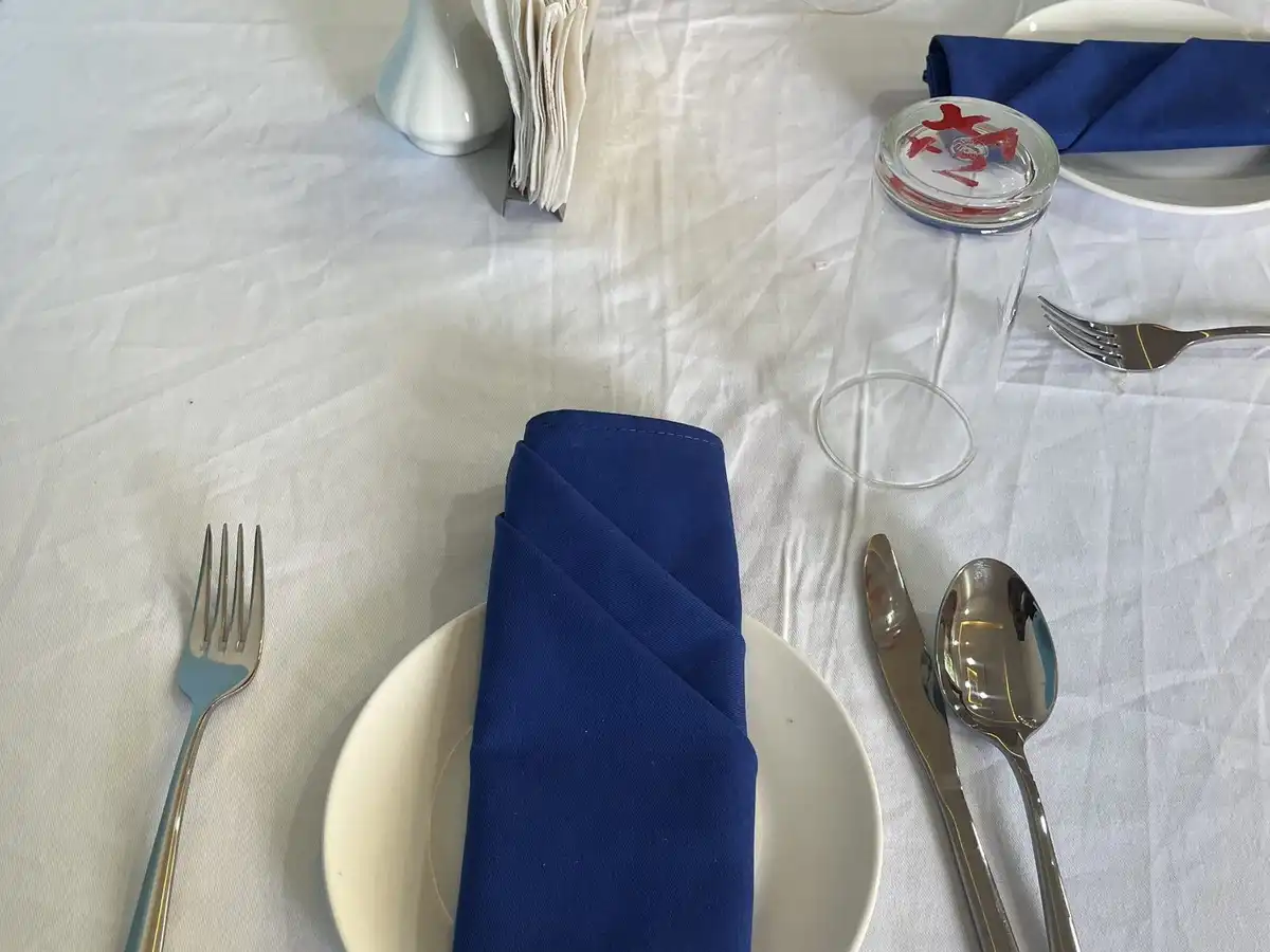 A place setting on a white tablecloth with a folded blue napkin, fork, knife and spoon