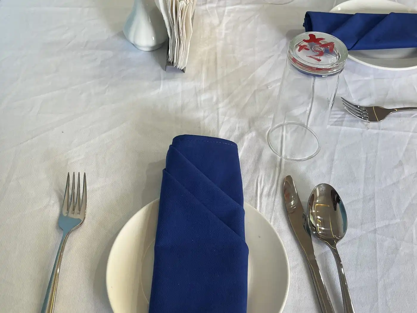 A place setting on a white tablecloth with a folded blue napkin, fork, knife and spoon