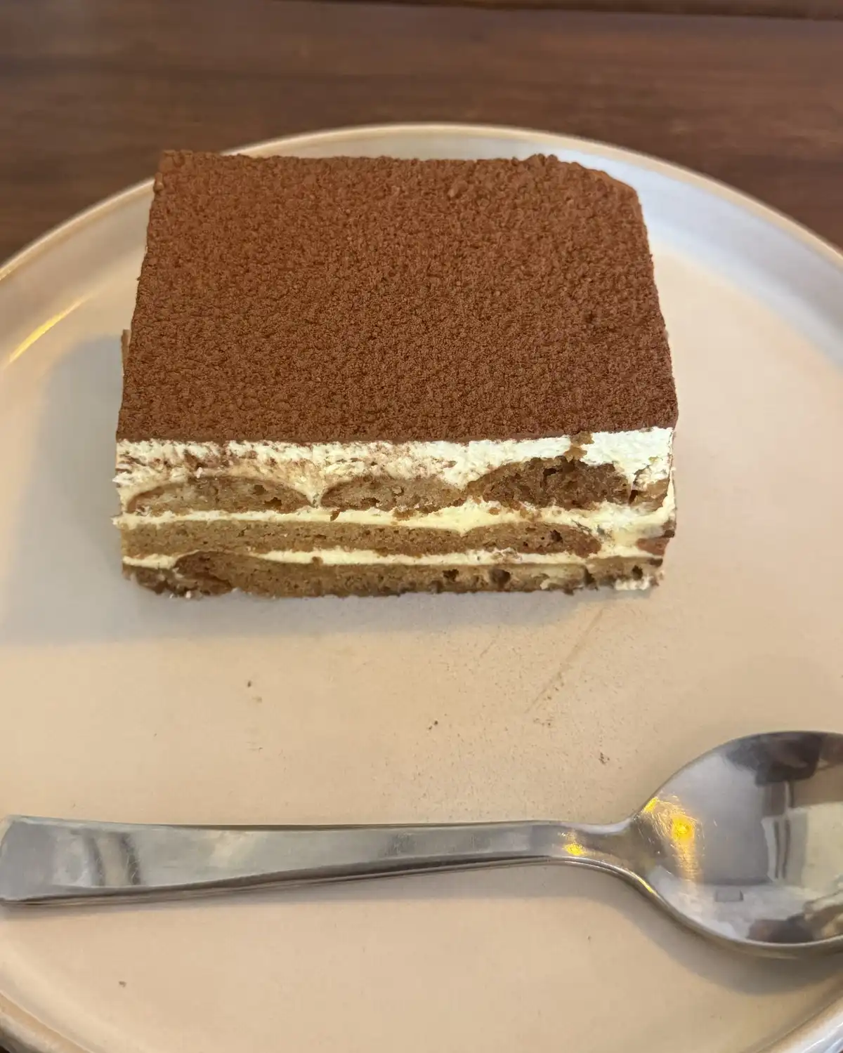 A square of tiramisu dusted with cocoa on a pale plate with a spoon